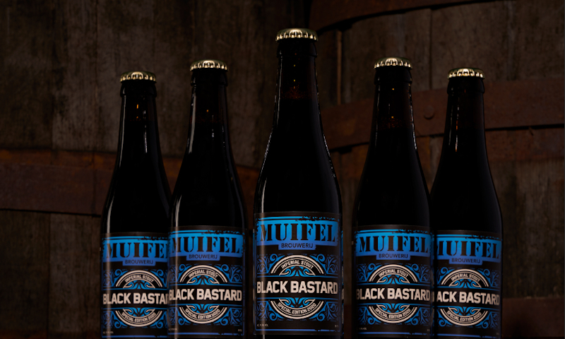 Vijf flessen Black Bastard van Muifelbrouwerij op een rij voor voor een paar houten vaten